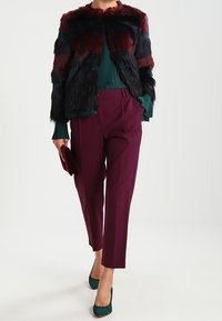 Faux fur jacket with striped dark red, green, and black design, paired with a green ribbed top and burgundy trousers; green heels.