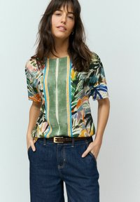 Blouse à manches courtes présentant un motif floral coloré, des accents verts et bleus, et un col rectangulaire, associée à un pantalon en denim foncé.