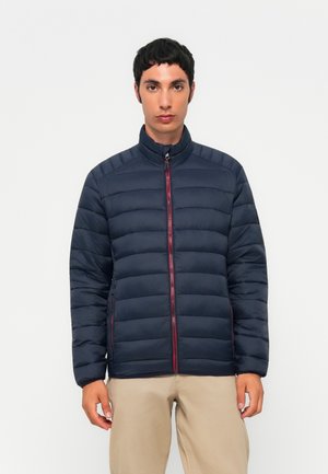 Muž nosící tmavě modrou prošívanou bundu s červeným zipem a béžové kalhoty, stojící před jednobarevným bílým pozadím.