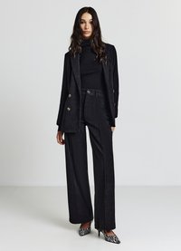 Schwarzer Cord-Anzug mit einem doppelt geknöpften Sakko und Hosen mit weitem Bein. Verfügt über glänzende goldene Knöpfe und kontrastierende gestreifte Stiletto-Absätze.