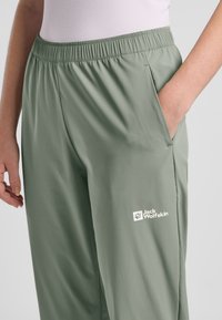 Lichtgroene sportbroek met een elastische tailleband, zijzakken en een wit "Jack Wolfskin"-logo nabij de onderkant. Glad textuur.