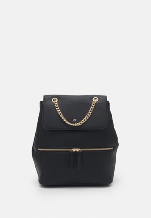 Sac à dos en cuir noir avec une poche zippée à l'avant et une poignée chaîne dorée sur le rabat supérieur, sur un fond clair uni.
