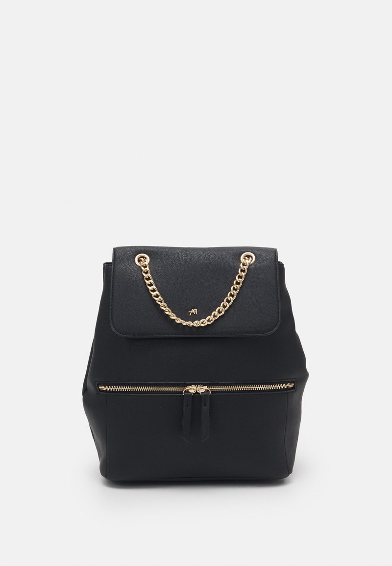 Sac à dos en cuir noir avec une poche zippée à l'avant et une poignée chaîne dorée sur le rabat supérieur, sur un fond clair uni.