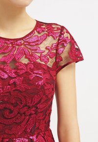 Robe rouge en sequins avec broderies florales, manches en maille transparente et col rond. Présente un motif texturé sur une base unie.