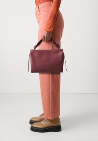 Burgundy bőr kézitáska fonott fogantyúval, világos rózsaszín, bővített lábú nadrággal és bézs fűzős csizmával párosítva. Sima anyag és minimális részletesség jellemzi.