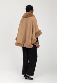 Poncho marrone con bordino in pelliccia, dotato di colletto e design ampio. Abbinato a pantaloni neri a gamba larga e scarpe nere lucide.