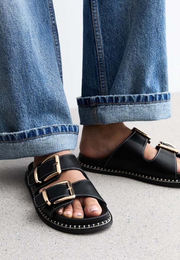 Pieds portant des sandales en cuir noir avec deux grandes boucles dorées, associés à un jean denim bleu roulé aux poignets sur un sol gris.