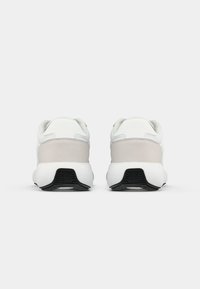 Paire de baskets blanches et gris clair vues de dos avec des semelles blanches épaisses et une bande de roulement noire sur fond blanc.