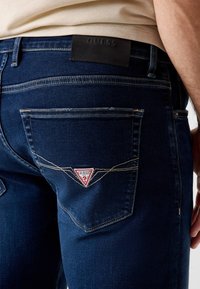 Vista posteriore di una persona che indossa jeans Guess blu scuro con il logo triangolare sulla tasca posteriore e una toppa nera del marchio in vita.