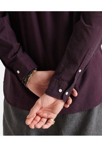 Persona con le mani incrociate dietro la schiena, indossante una camicia a maniche lunghe di un viola scuro con bottoni bianchi e pantaloni grigi, accessoriate con braccialetti e un anello.