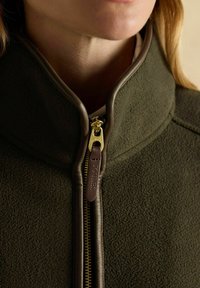 Zbliżenie osoby w oliwkowej polarowej kurtce z wysokim kołnierzem i złotym zamkiem błyskawicznym z brązowym skórzanym uchwytem.