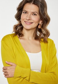 Lange gele cardigan van textuurgebreid materiaal, met een V-hals en geribde boorden, gedragen over een wit topje. Model met licht golvend haar.