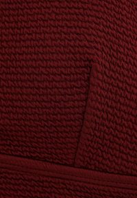 Bordo tkanina s teksturiranim, skupljenim uzorkom pletenine, blago presavijena pri dnu s horizontalnom linijom šava.