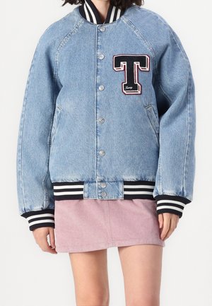 Kvinde iført en oversized lyseblå denim varsity-jakke med sort-hvide stribede manchetter og krave, kombineret med et lyserødt fløjlsnederdel.
