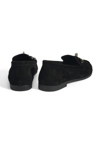 Zwarte suede loafers met een gladde textuur, een platte rubberen zool en een decoratieve metalen accent op het bovenwerk. De achterkant toont de ontwerpelementen.