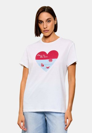 Donna che indossa una t-shirt bianca con un cuore rosso con conchiglie, un granchio e il testo "Amore al sole", abbinata a jeans azzurri chiari.