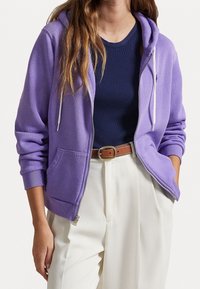 Felpa viola con zip e tasca frontale, con coulisse bianche e polsini a coste, indossata sopra una maglietta a coste blu navy e pantaloni bianchi.