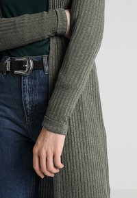 Cardigan grigio a coste indossato sopra una maglietta verde scuro, con maniche lunghe e una vestibilità ampia. Sono visibili dei jeans di jeans e una cintura nera con hardware in argento.