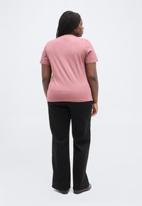 Donna con i capelli intrecciati che indossa una t-shirt rosa, pantaloni neri e scarpe, in piedi di spalle su uno sfondo bianco uniforme.