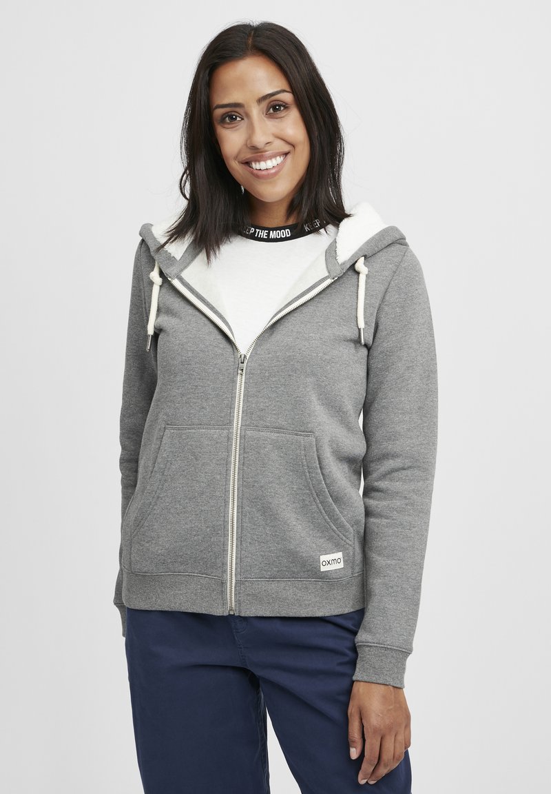 Gray zip-up hoodie with a white-fleece lined hood, drawstrings, and front pockets. Features a logo tag at the hem and ribbed cuffs.