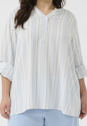 Femme portant un chemisier blanc ample avec des rayures verticales bleu clair et trois boutons, associé à un pantalon bleu clair, les bras détendus sur les côtés.