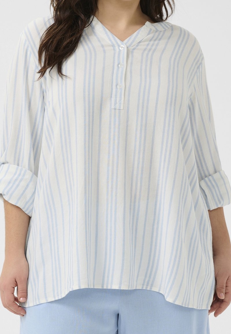 Femme portant un chemisier blanc ample avec des rayures verticales bleu clair et trois boutons, associé à un pantalon bleu clair, les bras détendus sur les côtés.