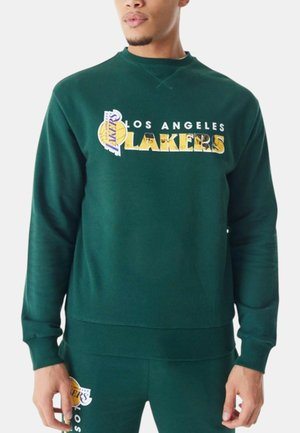 Uomo che indossa una felpa verde scuro dei Los Angeles Lakers e pantaloni abbinati con loghi della squadra e testo sul petto e sulla coscia.