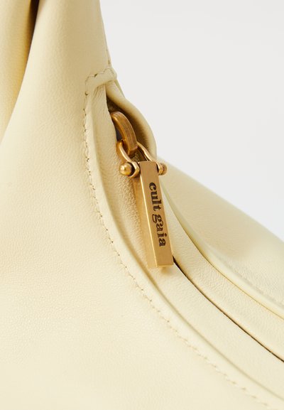Cream leather handbag featuring a curved design, gold hardware, a zipper closure, and a metallic logo tag with "cult gaia" engraved.