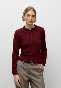 Woman wearing maroon long-sleeve polo shirt, plaid pants with black belt, and large metallic earrings, standing against plain background.