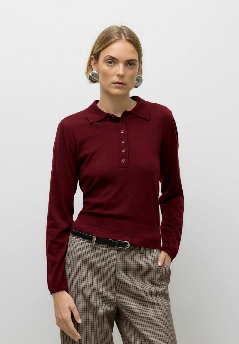Woman wearing maroon long-sleeve polo shirt, plaid pants with black belt, and large metallic earrings, standing against plain background.