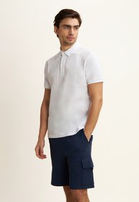 Polo shirt bianco a maniche corte con collo a tre bottoni, abbinato a pantaloni cargo blu scuro con tasca laterale e dettaglio del logo.