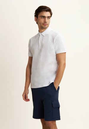 Polo shirt bianco a maniche corte con collo a tre bottoni, abbinato a pantaloni cargo blu scuro con tasca laterale e dettaglio del logo.