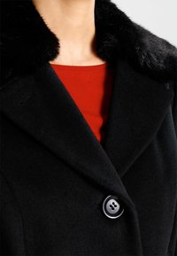 Zwarte wollen jas met grote knoop en zachte zwarte bontkraag, gedragen over een rode ronde hals top, waarbij de nek en bovenste gedeelte van de borst zichtbaar zijn.