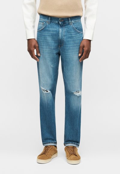 Άντρας που φοράει ανοιχτό μπλε φθαρμένα τζιν, μπεζ πουλόβερ, λευκό πουκάμισο και καφέ σουέτ αθλητικά παπούτσια, στέκεται μπροστά σε απλό φόντο.