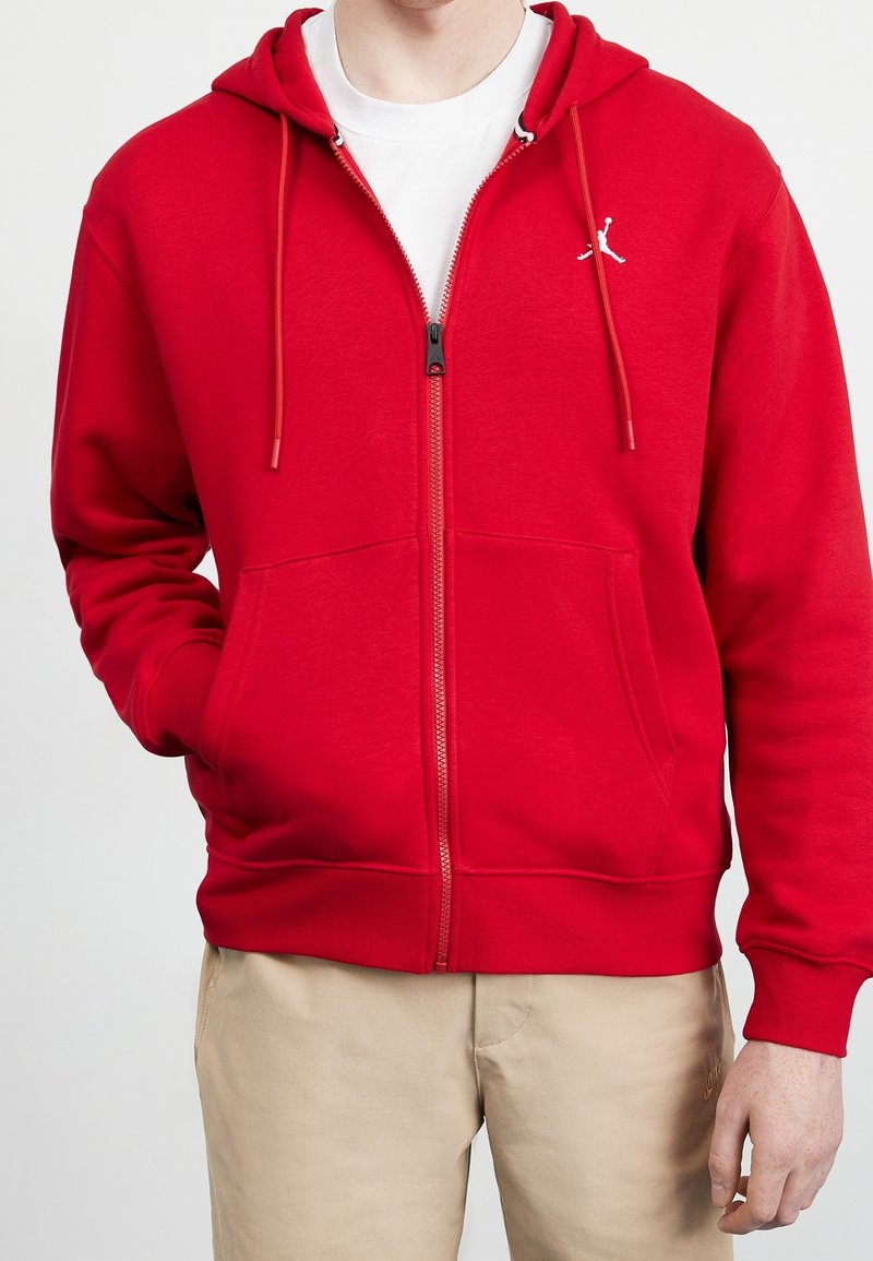 Man in een rode rits-hoodie met een voorzak en beige broek, met één hand in de zak, staande tegen een effen achtergrond.