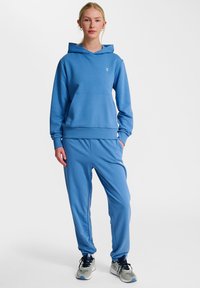 Blå huvtröja med en framficka och huva, tillsammans med matchande blå joggingsbyxor. Båda i mjukt tyg och med en diskret logotypdetalj.