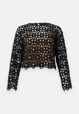 Haut long à manches longues en crochet noir avec motif floral et petites garnitures de miroirs ronds sur un fond clair uni.