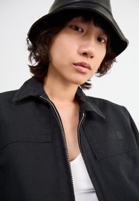 Must värvi kangast jakki, millel on lukuga esikülg, terav krae ja kaks rinna taskut. Seljas on valge ribidega tank top. Näha on must bucket hat.