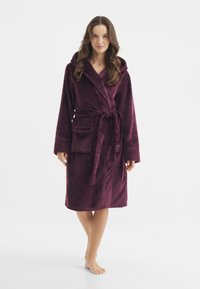 Accappatoio in pile con cappuccio di un profondo bordeaux, con maniche lunghe, cintura da annodare e una tasca applicata sul lato sinistro, realizzato in un tessuto morbido.