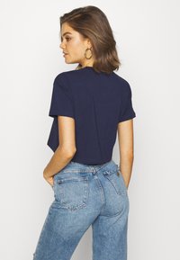 Mujer con cabello hasta los hombros que lleva pendientes de aro, una camiseta corta azul marino y vaqueros de tiro alto de color azul claro, de pie con las manos en los bolsillos.