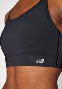 Close-up de uma pessoa a usar um top desportivo preto com o logótipo da New Balance na faixa sob o peito, a mostrar um abdómen tonificado.