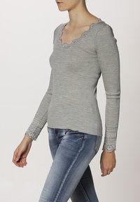 Grå ribbad långärmad topp med en vågig halsringning och spetsdetaljer vid ärmsluten, kombinerad med blå jeans.