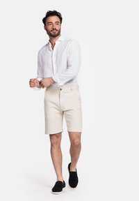 Wit linnen overhemd met knopen, beige shorts en zwarte instapschoenen. Model staat en past de manchetten van het overhemd aan, met zichtbare korte mouwen.