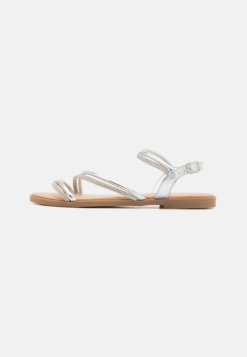 Silver flat sandals with crisscross straps, rhinestone accents, and a brown sole. Adjustable ankle strap for secure fit.
