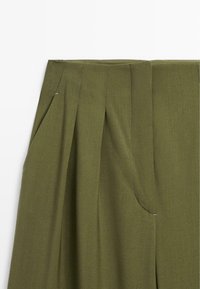 Pantalons vert olive avec des plis, une texture lisse, des poches latérales et des détails cousus autour de la taille et des poches.
