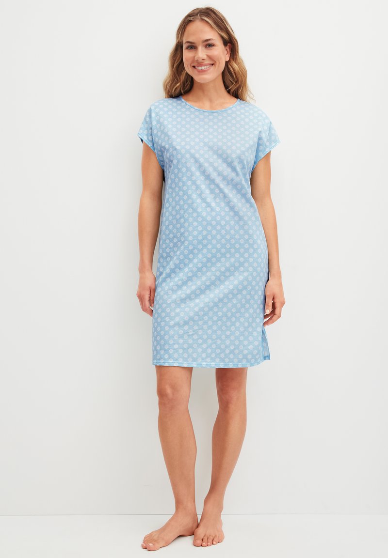 Femme debout pieds nus portant une robe bleu clair mi-genoux à pois blancs, manches courtes et fentes latérales, souriant à la caméra.