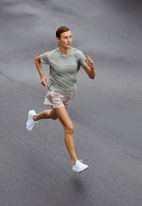 Femme en chemise grise et short beige courant sur un pavé gris lisse, capturée en plein pas avec les deux pieds en l'air.