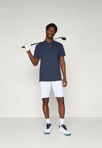 Tengerészkék, rövid ujjú pólóing álló gallérral, elöl cipzárral, fehér sorttal, fehér és tengerészkék golfcipőt viselve, valamint golfütőt tartva.