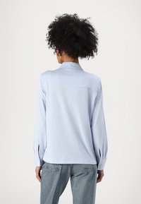 Woman with curly hair wearing a light blue long-sleeve blouse and blue denim jeans, facing away from the camera against a plain background.
