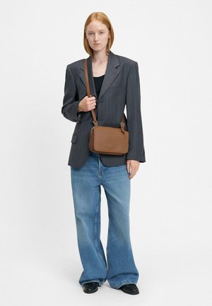 Braune Leder Umhängetasche in rechteckiger Form, mit dezenten Nähten und einem verstellbaren Riemen. Das Model trägt ein dunkles Blazer und ausgestellte Jeans.